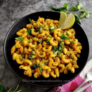 masala macaroni served in a black bowl and garnished with cilantro and lemon wedges