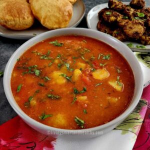 aloo tamatar ki sabji served in a bowl with bedmi pooris on a plate kept on the top left side