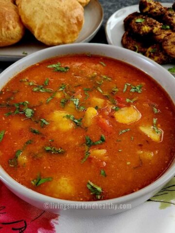 aloo tamatar ki sabji served in a bowl with bedmi pooris on a plate kept on the top left side