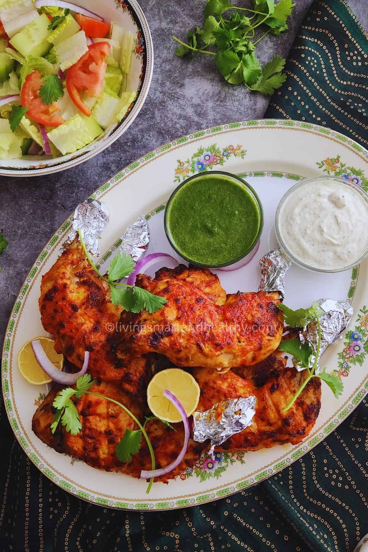 Oven baked tandoori chicken leg quarters on a serving platter with mint chutney, white sauce, and salad