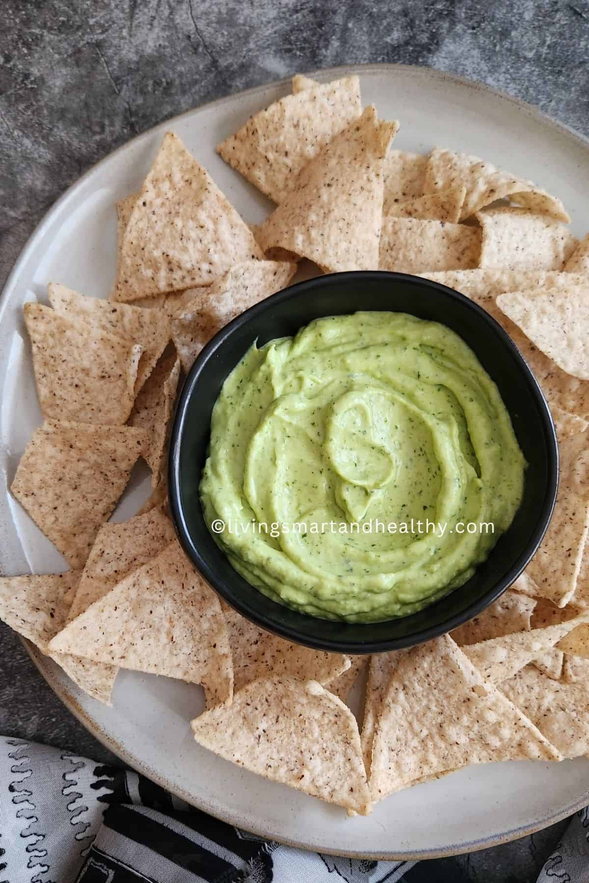 Bowl of smooth green avocado dip with Greek yogurt surrounded by tortilla chips, pita bread, and fresh cut vegetables on a rustic table